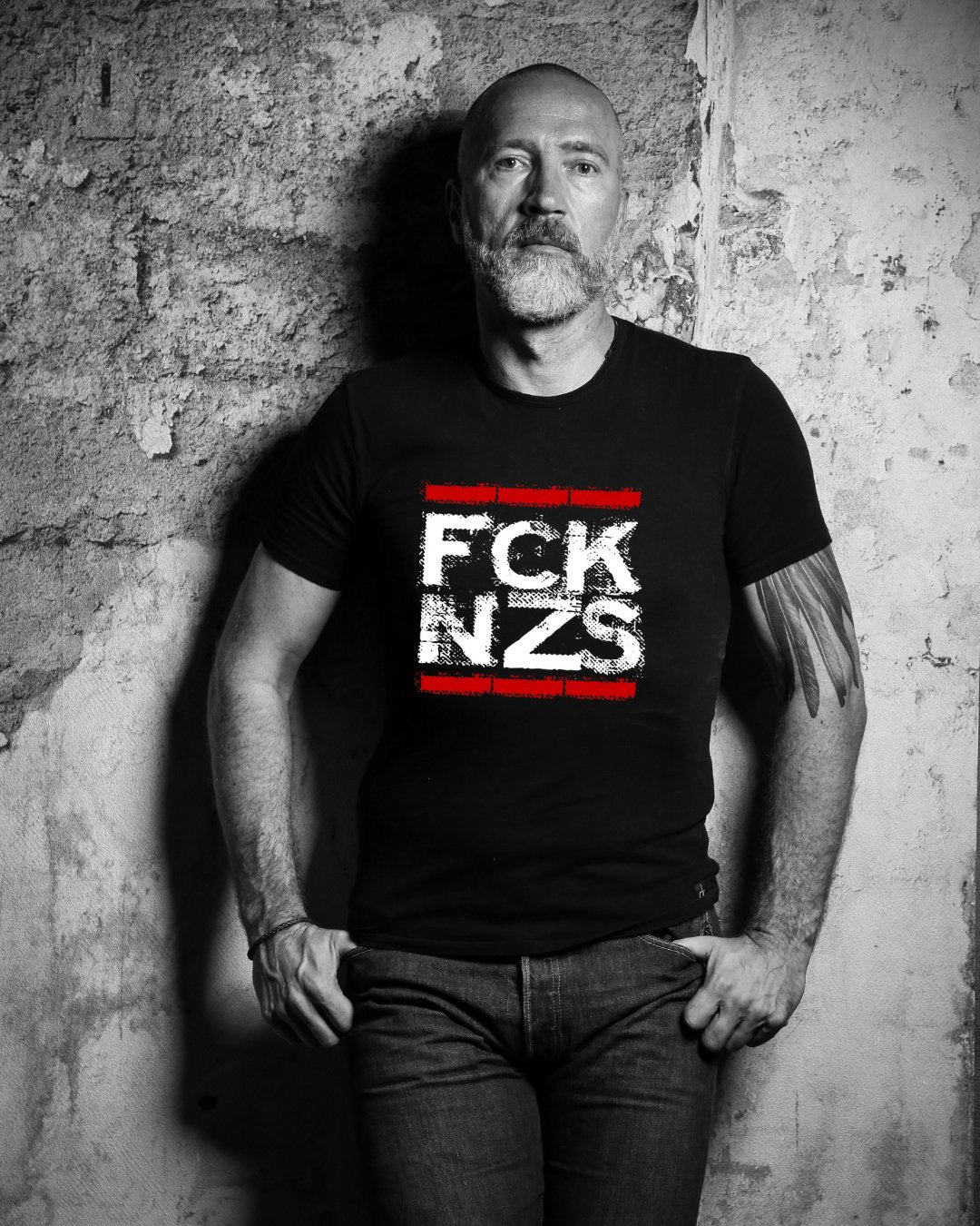 „Mann vor rauer Wand in schwarz-weißem Foto, trägt schwarzes T-Shirt mit weiß-rotem Statement: FCK NZS – ein klares Zeichen gegen Faschismus und rechte Hetze.“