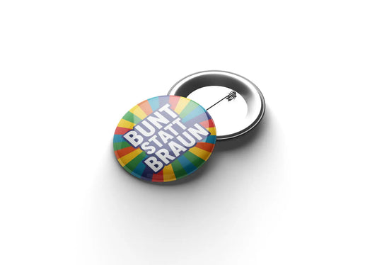 „Runder Button mit farbigem, strahlenförmigem Hintergrund in Rot, Gelb, Grün, Blau und Orange; in der Mitte steht auf dunkelblauem Kreis die weiße Aufschrift ‚BUNT STATT BRAUN‘.“