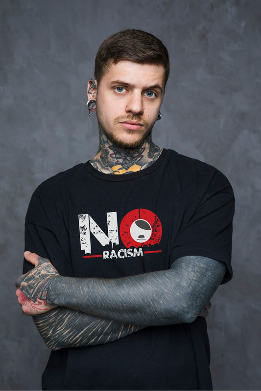 Person trägt schwarzes Unisex T‑Shirt mit großem NO RACISM Frontprint und Ärmeldruck, Arme verschränkt.