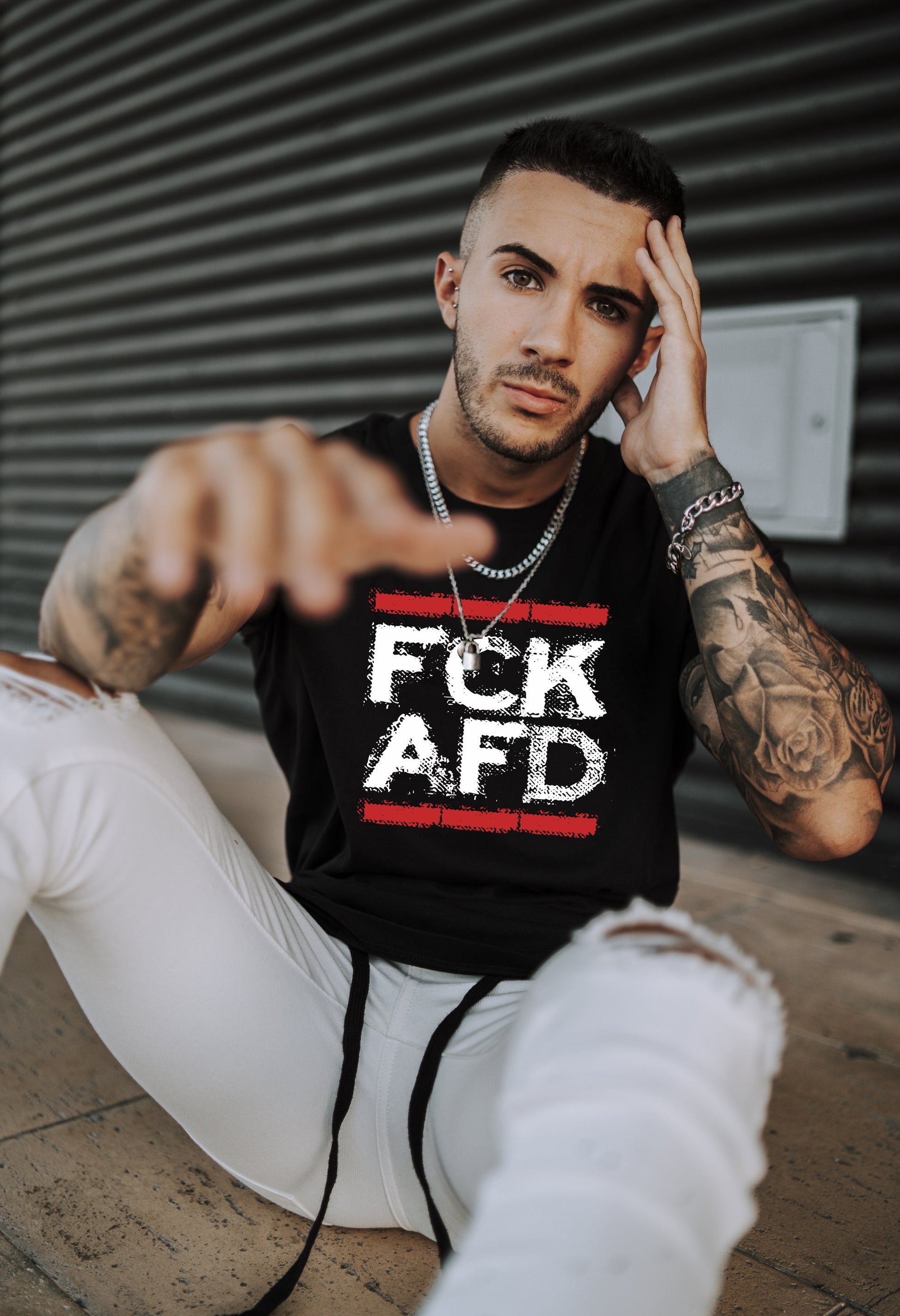 „Person trägt ein schwarzes ärmelloses Shirt mit weiß‑rotem FCK AFD Frontprint zwischen roten Balken, kombiniert mit weißen Jeans und Tattoos.“


