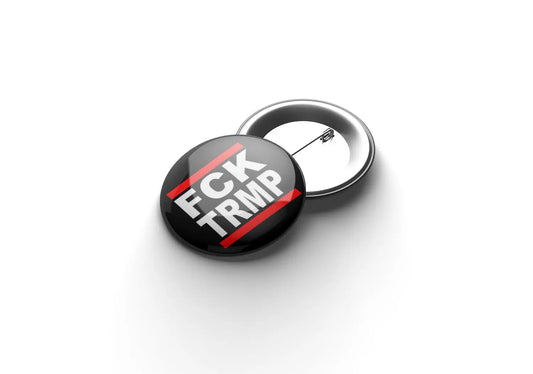 „Runder Button mit der Aufschrift ‚FCK TRMP‘ in weißer Blockschrift auf rotem Hintergrund; daneben ein zweiter Button mit sichtbarer Metallrückseite und Sicherheitsnadel.“