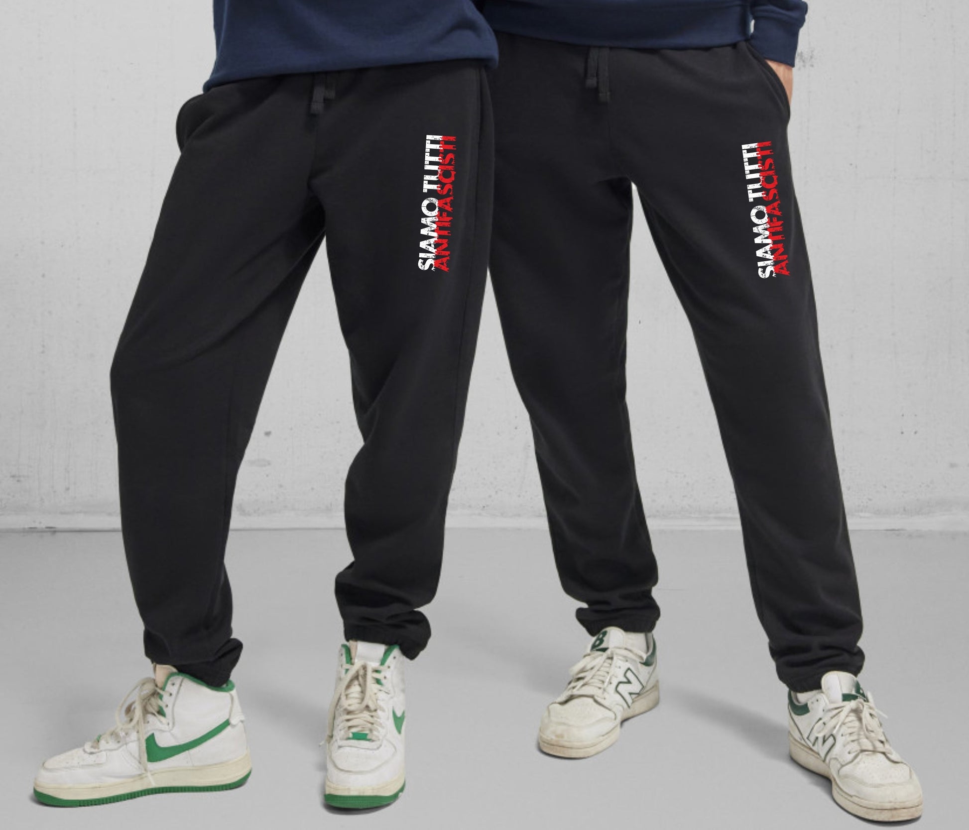 Zwei Personen tragen schwarze Bio-Jogginghosen mit vertikalem Schriftzug „SIAMO TUTTI ANTIFASCISTI“ auf dem Oberschenkel. Die Worte „SIAMO TUTTI“ sind weiß, „ANTIFASCISTI“ erscheint in rotem Druck. Beide stehen nebeneinander vor grauem Hintergrund und tragen weiße Sneaker. Das Design vermittelt eine klare antifaschistische Botschaft im sportlich-urbanen Stil.