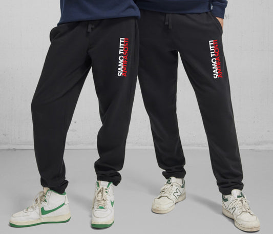 Zwei Personen tragen schwarze Bio-Jogginghosen mit vertikalem Schriftzug „SIAMO TUTTI ANTIFASCISTI“ auf dem Oberschenkel. Die Worte „SIAMO TUTTI“ sind weiß, „ANTIFASCISTI“ erscheint in rotem Druck. Beide stehen nebeneinander vor grauem Hintergrund und tragen weiße Sneaker. Das Design vermittelt eine klare antifaschistische Botschaft im sportlich-urbanen Stil.