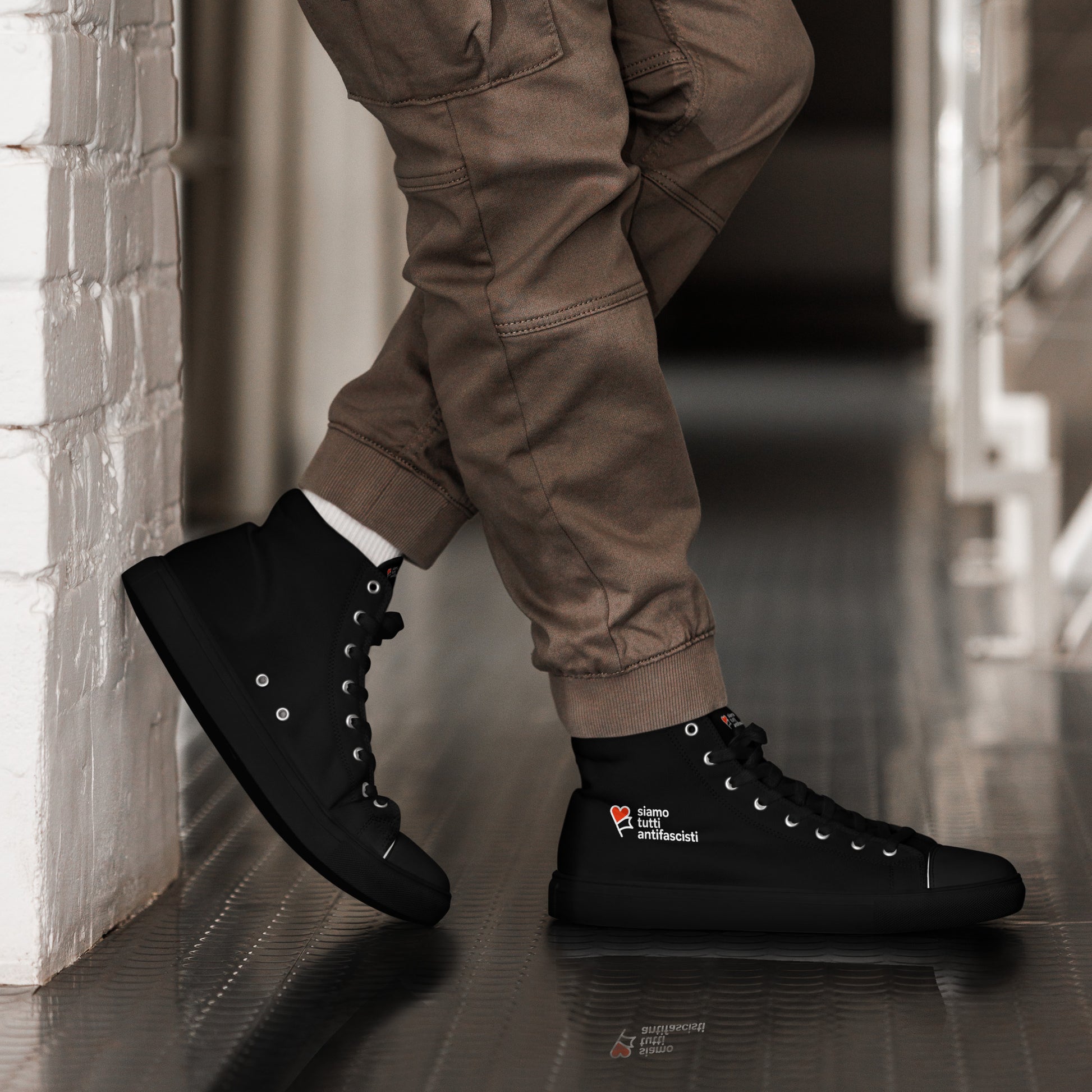 „Schwarze High‑Top‑Sneaker mit weißer Sohle und weißen Schnürsenkeln. Auf der Außenseite befindet sich ein rotes Herz mit dem italienischen Schriftzug ‚siamo tutti antifascisti‘ als politisches Statement.“